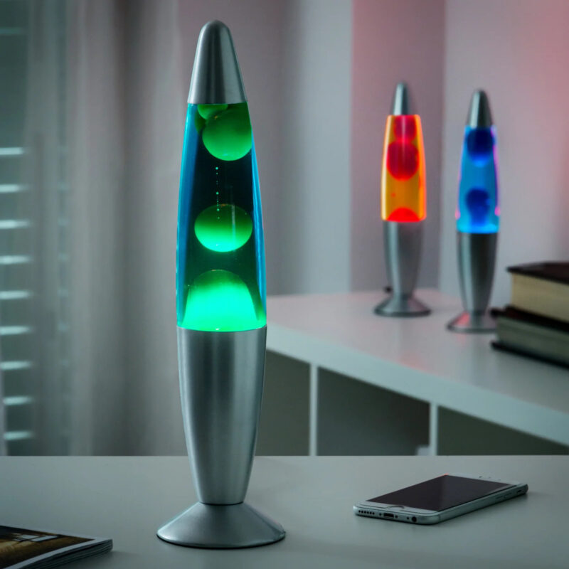Лава лампа InnovaGoods с метална основа и зелено осветление, разположена на бюро до смартфон, използвана като декоративно осветление в модерен интериор.
